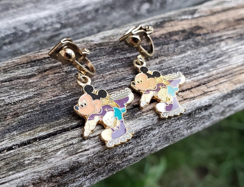 K&ouml;nnte beinhalten: Ein Paar goldfarbene Ohrclips mit Mickey Mouse und Minnie Mouse beim Rollschuhlaufen. Die Ohrringe sind mit buntem Email verziert und haben ein lila, gr&uuml;nes und gelbes Farbschema.