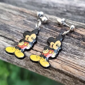 May include: A pair of Mickey Mouse dangle earrings. Each earring features a black silhouette of Mickey Mouse with yellow shoes, red shorts, and a yellow face. The earrings have silver-colored posts and connecting chains.