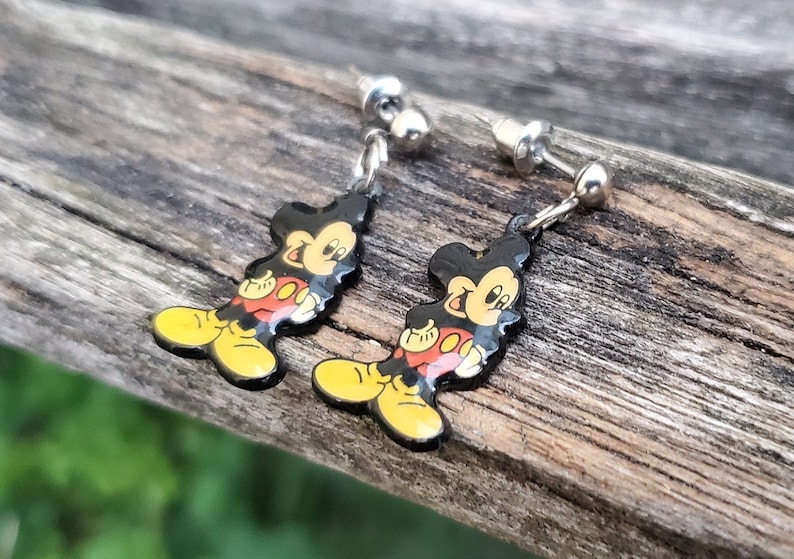 May include: A pair of Mickey Mouse dangle earrings. Each earring features a black silhouette of Mickey Mouse with yellow shoes, red shorts, and a yellow face. The earrings have silver-colored posts and connecting chains.