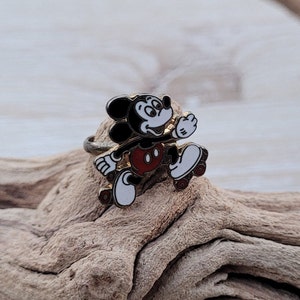 May include: A gold-tone ring featuring a black and white enamel Mickey Mouse character on roller skates. The ring is set on a piece of driftwood.