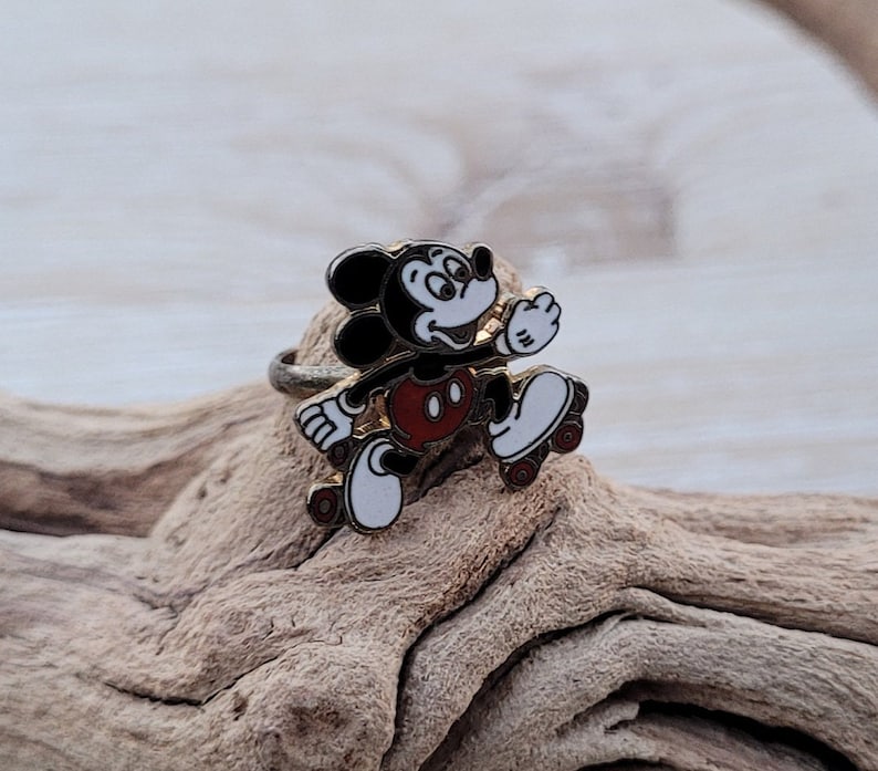 May include: A gold-tone ring featuring a black and white enamel Mickey Mouse character on roller skates. The ring is set on a piece of driftwood.