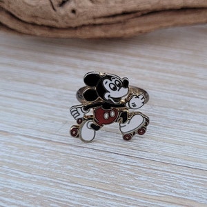 May include: A gold-toned ring featuring a cartoon image of Mickey Mouse roller skating. Mickey is wearing red shorts and white roller skates.