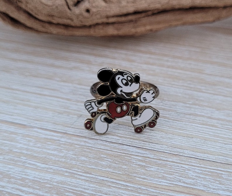 May include: A gold-toned ring featuring a cartoon image of Mickey Mouse roller skating. Mickey is wearing red shorts and white roller skates.