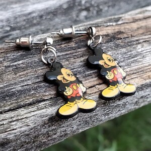 May include: A pair of Mickey Mouse earrings. Each earring features a black, red, yellow, and white cartoon figure of Mickey Mouse dangling from a silver post. The earrings are set against a weathered wooden surface.