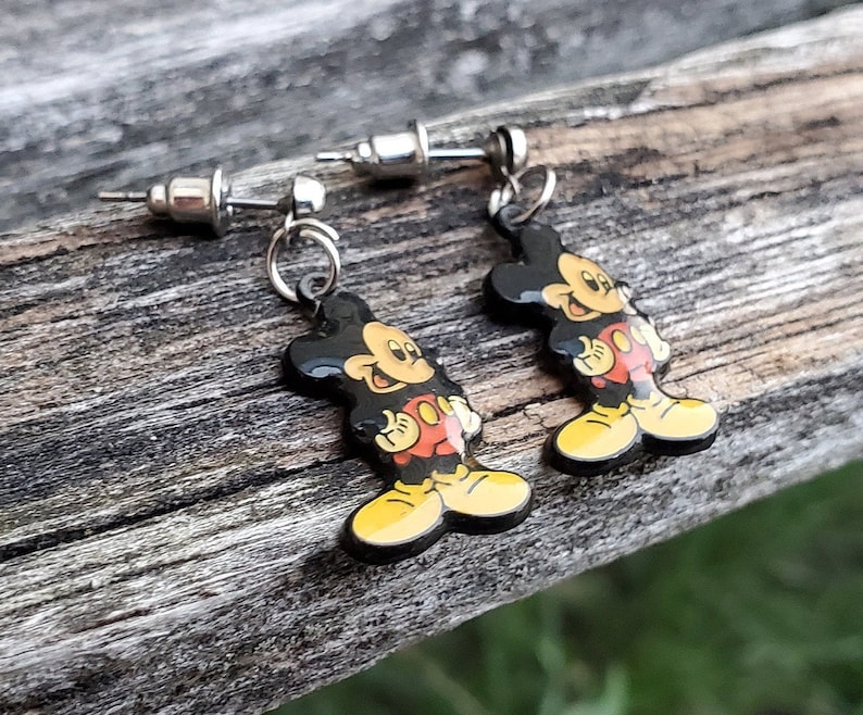 May include: A pair of Mickey Mouse earrings. Each earring features a black, red, yellow, and white cartoon figure of Mickey Mouse dangling from a silver post. The earrings are set against a weathered wooden surface.