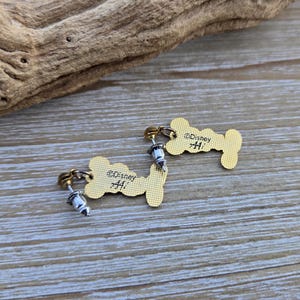 Puede incluir: Un par de pendientes de color dorado con forma de hueso de perro. Cada pendiente tiene un poste de plata y un peque&ntilde;o dije de plata. La parte posterior de cada pendiente est&aacute; grabada con el texto "&copy;Disney #41".