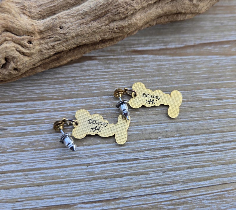 Puede incluir: Un par de pendientes de color dorado con forma de hueso de perro. Cada pendiente tiene un poste de plata y un peque&ntilde;o dije de plata. La parte posterior de cada pendiente est&aacute; grabada con el texto "&copy;Disney #41".