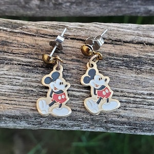 Puede incluir: Un par de pendientes de Mickey Mouse de color dorado con pantalones cortos rojos y zapatos negros. Los pendientes son de estilo colgante con un poste plateado.