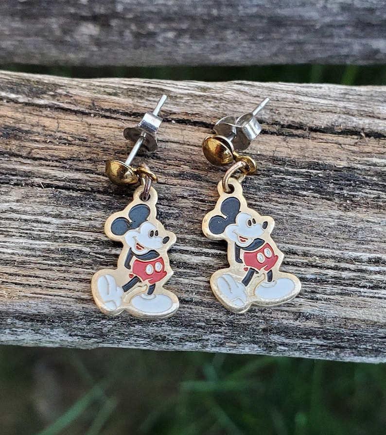Puede incluir: Un par de pendientes de Mickey Mouse de color dorado con pantalones cortos rojos y zapatos negros. Los pendientes son de estilo colgante con un poste plateado.