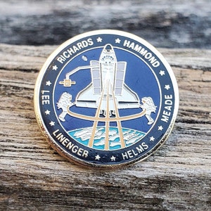 May include: A gold-colored pin with a black and blue design. The pin features a space shuttle, astronauts, and the names "Richards, Hammond, Meade, Linenger, Helms".