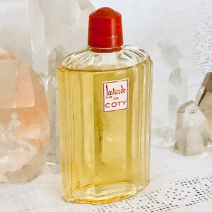 Pu&ograve; includere: Un flacone di profumo Coty L'avande in vetro trasparente con un tappo rosso. Il flacone &egrave; appoggiato su una superficie bianca con cristalli di quarzo trasparenti.