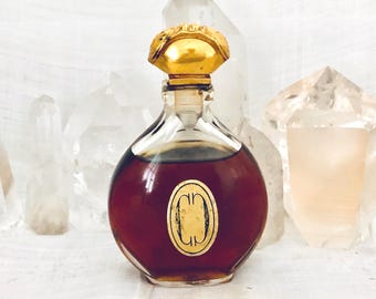 May include: A round glass perfume bottle filled with dark amber liquid, featuring a gold-colored stopper with a textured top and a central oval gold label displaying a stylized double C logo. The bottle has a clear neck and a wide, rounded base.