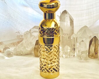 May include: A gold-colored perfume bottle with a decorative lattice design. The bottle has a rounded top and a matching cap. The bottle is displayed on a white surface with clear quartz crystals in the background.