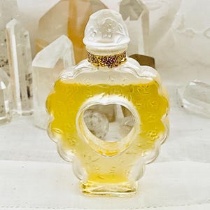 May include: A heart-shaped glass bottle of perfume with a white cap and gold accents. The bottle is filled with a light yellow liquid.