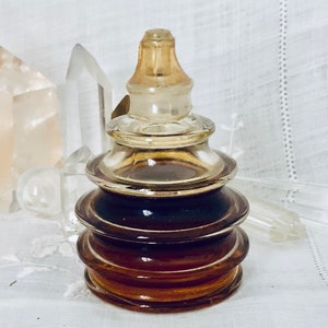 May include: A vintage glass perfume bottle with a layered, pagoda-like design. The bottle is filled with a dark brown liquid and has a clear glass stopper.