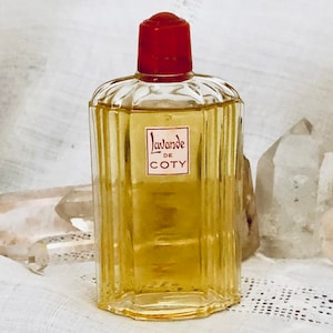 Pu&ograve; includere: Un flacone di profumo vintage in vetro di Lavande di Coty L'Oreal. Il flacone ha un tappo rosso ed &egrave; riempito con un liquido giallo chiaro. Il flacone &egrave; appoggiato su una superficie di tessuto bianco con cristalli trasparenti.