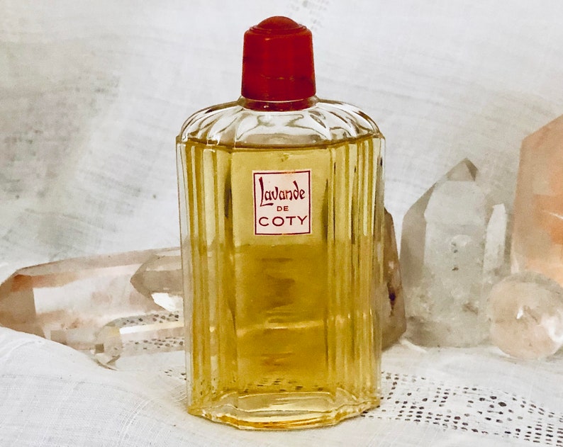 Pu&ograve; includere: Un flacone di profumo vintage in vetro di Lavande di Coty L'Oreal. Il flacone ha un tappo rosso ed &egrave; riempito con un liquido giallo chiaro. Il flacone &egrave; appoggiato su una superficie di tessuto bianco con cristalli trasparenti.