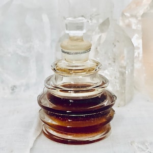 May include: A clear glass perfume bottle with a stacked, tiered design. The bottle is filled with a dark amber liquid and has a clear stopper. The bottle is set against a soft, white background, with crystals in the background.