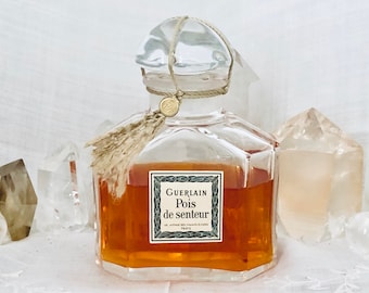 Puede incluir: Un frasco de perfume vintage de vidrio de Guerlain Pois de Senteur con un borla marrón y una tapa dorada. El frasco está lleno de un líquido dorado y está sentado sobre una superficie blanca con dos cristales de cuarzo transparentes.