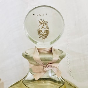K&ouml;nnte beinhalten: Eine Vintage-Glasflasche mit einem durchsichtigen Glasstopfen, der mit einer goldenen Krone und einem Wappen verziert ist. Die Flasche ist mit einem rosa Band gebunden.
