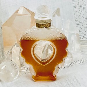 May include: A clear glass bottle with a heart-shaped opening, filled with a golden liquid. The bottle is decorated with floral designs and has a gold-colored stopper.