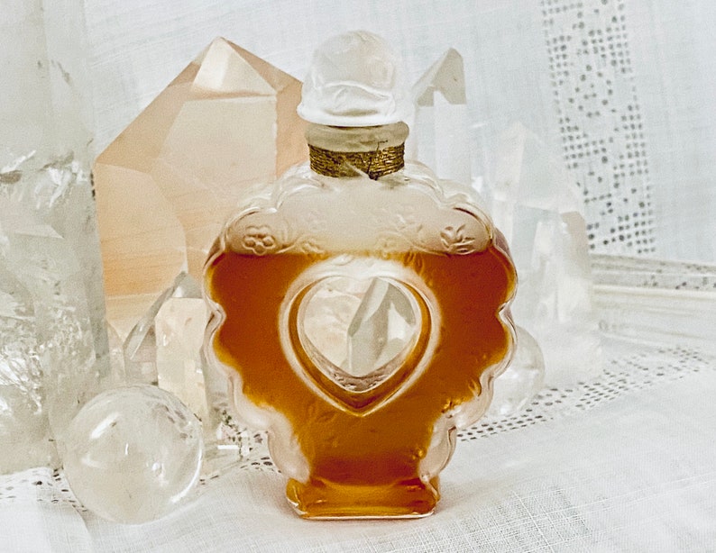 May include: A clear glass bottle with a heart-shaped opening, filled with a golden liquid. The bottle is decorated with floral designs and has a gold-colored stopper.