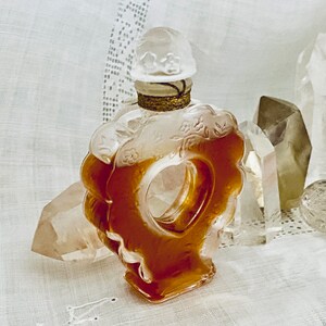 May include: A vintage glass perfume bottle with a floral design and a clear, amber-colored liquid. The bottle is shaped like a heart and has a decorative stopper. The bottle is sitting on a white fabric surface.