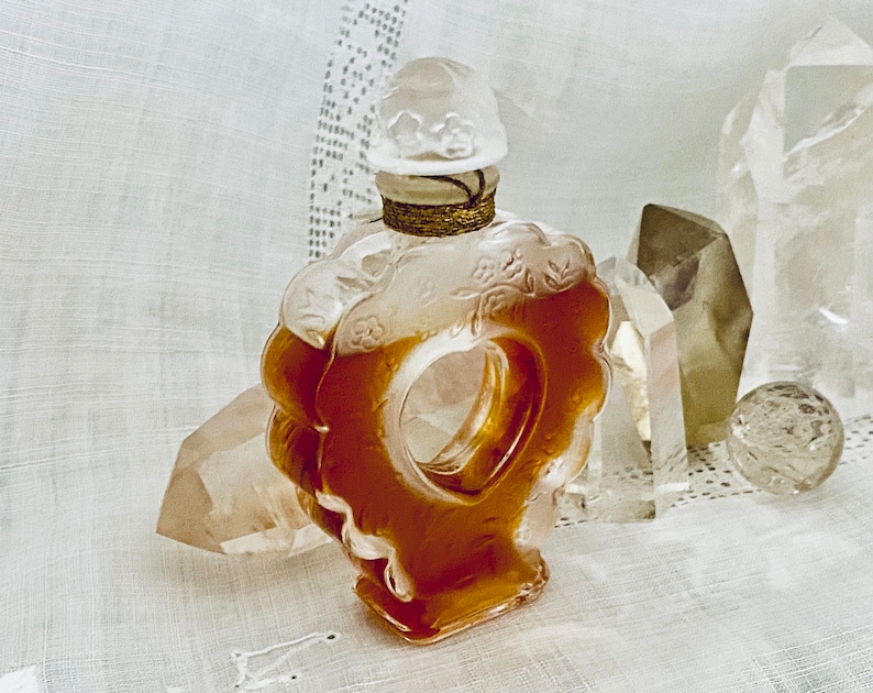 May include: A vintage glass perfume bottle with a floral design and a clear, amber-colored liquid. The bottle is shaped like a heart and has a decorative stopper. The bottle is sitting on a white fabric surface.