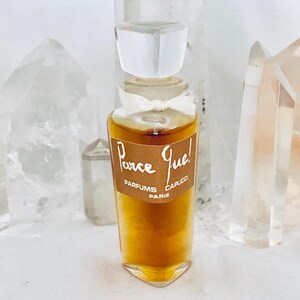 May include: A clear glass perfume bottle with a tapered base and a faceted clear stopper. A white ribbon is tied around the neck. The label is brown with white text that reads Parce Que! PARFUMS CAPUCCI PARIS. The liquid inside is amber colored.