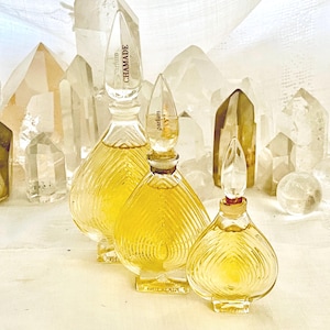 May include: Three vintage perfume bottles with clear, teardrop-shaped stoppers and a ribbed glass design. The bottles are filled with a golden liquid and are labeled "CHAMADE" and "parfum". The background features clear quartz crystals.