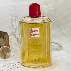 Pu&ograve; includere: Un flacone di profumo Coty L'avande in vetro trasparente con un tappo rosso. Il flacone &egrave; appoggiato su una superficie bianca.