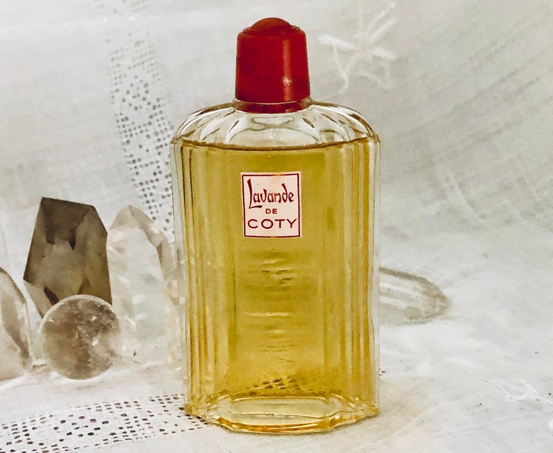 Pu&ograve; includere: Un flacone di profumo Coty L'avande in vetro trasparente con un tappo rosso. Il flacone &egrave; appoggiato su una superficie bianca.