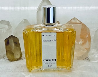 May include: A rectangular glass bottle of Caron Narcisse Noir Eau de Toilette. The bottle has a silver cap and contains a golden-colored liquid. The label reads "Caron Paris-France 85°".