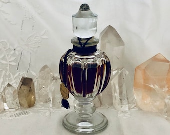 May include: A clear glass bottle with a textured surface and a black stopper. The bottle is filled with a dark brown liquid. The bottle is sitting on a white surface with several clear and pink crystals.