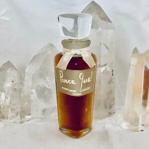 May include: A vintage glass bottle of "Ponce Gue!" perfume by Carucci Paris. The bottle is filled with a dark amber liquid and has a clear glass stopper. The bottle is sitting on a white surface.