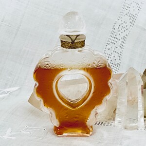 May include: A clear glass perfume bottle shaped like a heart with a gold stopper. The bottle is filled with a golden liquid and is sitting on a white surface.