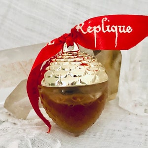 May include: A gold acorn-shaped ornament with a red ribbon tied around it. The ribbon has the word "Replique" written on it in white.