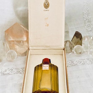 Pu&ograve; includere: Un flacone vintage di profumo Coty L'Aimant in una scatola bianca. Il flacone &egrave; in vetro trasparente con un tappo rosso e lettere dorate. La scatola ha una corona dorata e la parola "Coty" stampata su di essa.