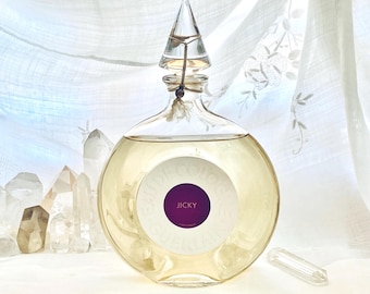 May include: A round glass perfume bottle with a clear, pointed stopper. The bottle contains a light yellow liquid and has a white label with the word "Jicky" in a purple circle. The bottle is surrounded by clear quartz crystals.