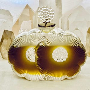 May include: A clear glass perfume bottle with a textured, floral design. The bottle is filled with a golden liquid and has a decorative stopper.