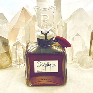 May include: A square amber-colored perfume bottle with a clear glass stopper featuring the letter "R". The bottle is labeled "Réplique PARIS". A red wax seal is attached to the bottle. The background features several clear quartz crystals.