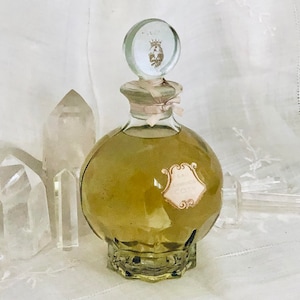 K&ouml;nnte beinhalten: Eine durchsichtige Glasflasche mit einem goldenen Verschluss und einem Etikett mit der Aufschrift "Eau de Cologne" in einer dekorativen Schriftart. Die Flasche ist mit einer hellgr&uuml;nen Fl&uuml;ssigkeit gef&uuml;llt und steht auf einer wei&szlig;en Oberfl&auml;che.