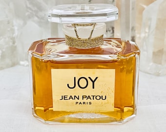 May include: A clear glass bottle of Joy perfume by Jean Patou Paris. The bottle is filled with a golden liquid and has a gold-coloured cap.