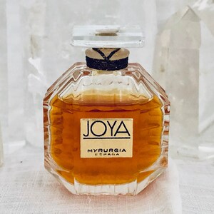 May include: A small, clear glass bottle of perfume with a gold label that reads "JOYA MYRURGIA ESPAÑA". The bottle is filled with a golden liquid.