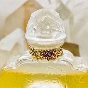 May include: A clear glass bottle with a gold-colored cap and a white floral design. The bottle is filled with a yellow liquid.