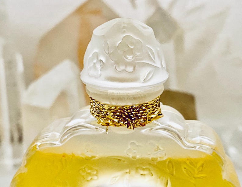 May include: A clear glass bottle with a gold-colored cap and a white floral design. The bottle is filled with a yellow liquid.