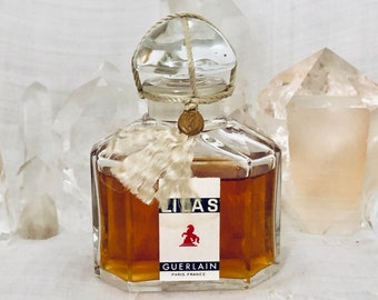 May include: A vintage glass bottle of Guerlain Lilas perfume. The bottle has a clear glass stopper with a white tassel and a gold seal. The label is white with black text and a red horse logo.