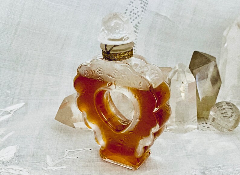 May include: A clear glass bottle with a heart-shaped design and a white stopper. The bottle is filled with a golden liquid and is surrounded by clear crystals.