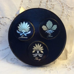 May include: A black round compact with three circular compartments, each featuring a different floral design and the name of the scent: Jasmine, Rose, and Ylang.