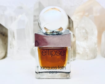 May include: A square glass perfume bottle containing amber liquid with the text ellipse jacques fath paris printed on the front. The bottle features a silver-toned rectangular cap topped with a clear, spherical glass orb ornament.
